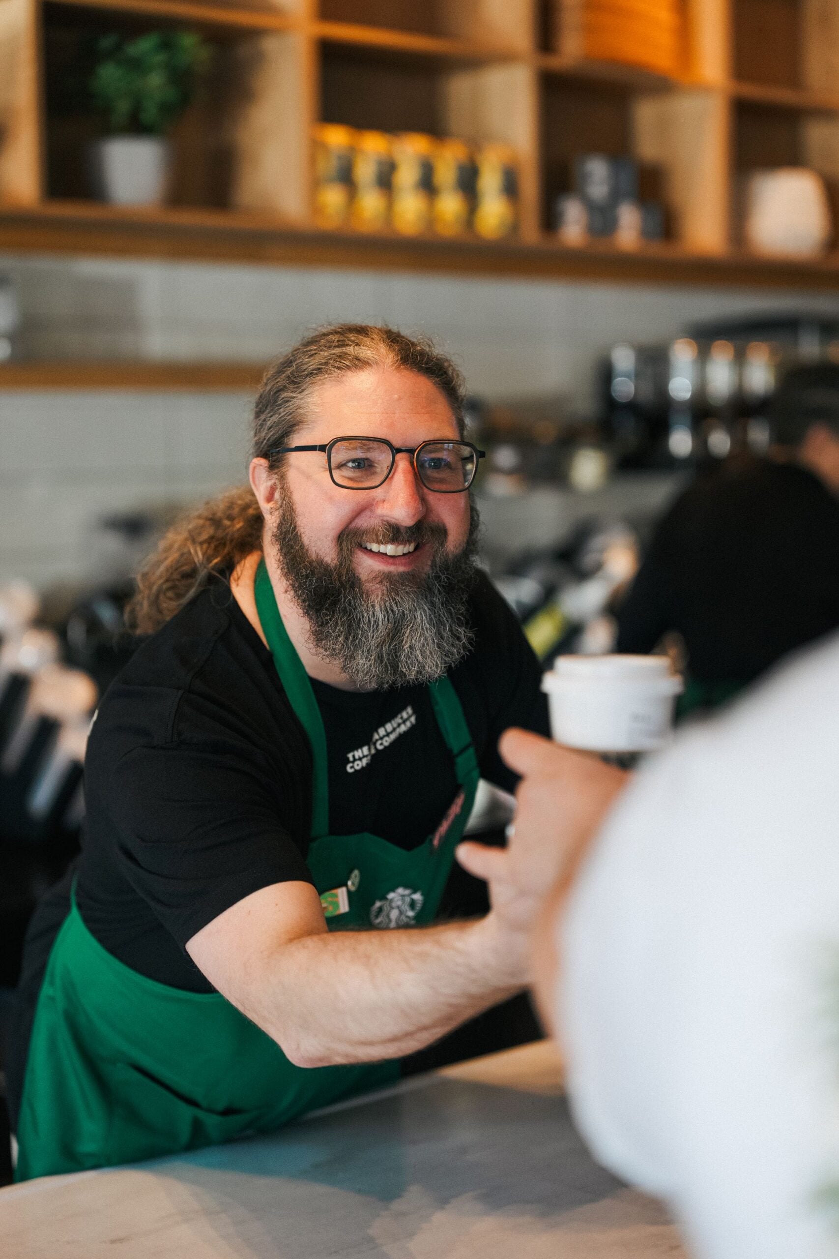 A smiling barista with long hair and a beard, wearing a green apron, hands a coffee cup to a customer at a café.