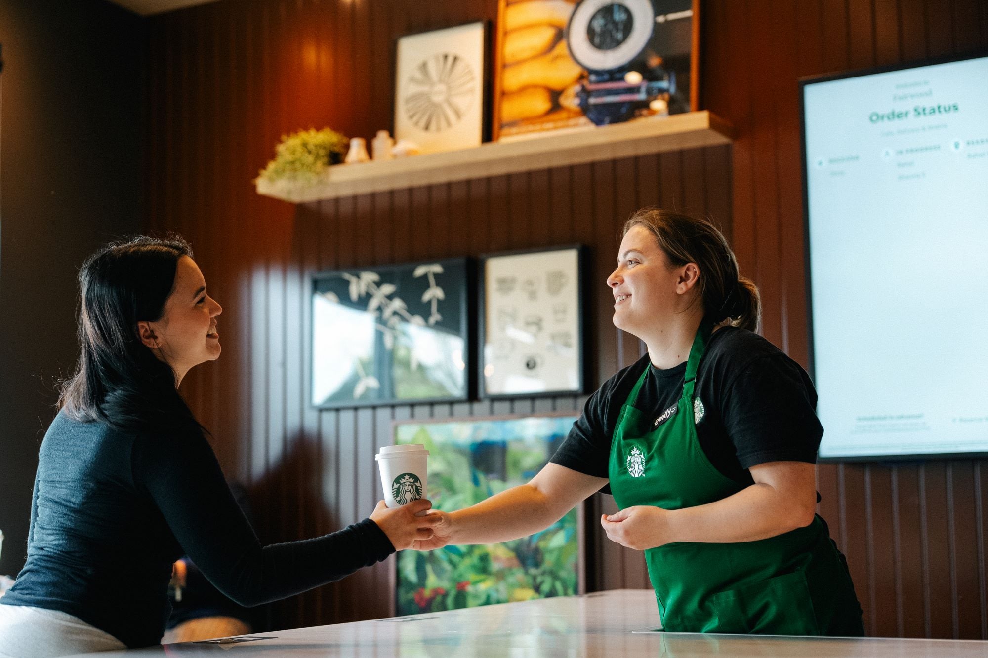 A barista in a green apron hands a coffee cup to a customer at a Starbucks counter. The setting features wooden paneling and decorative artwork.