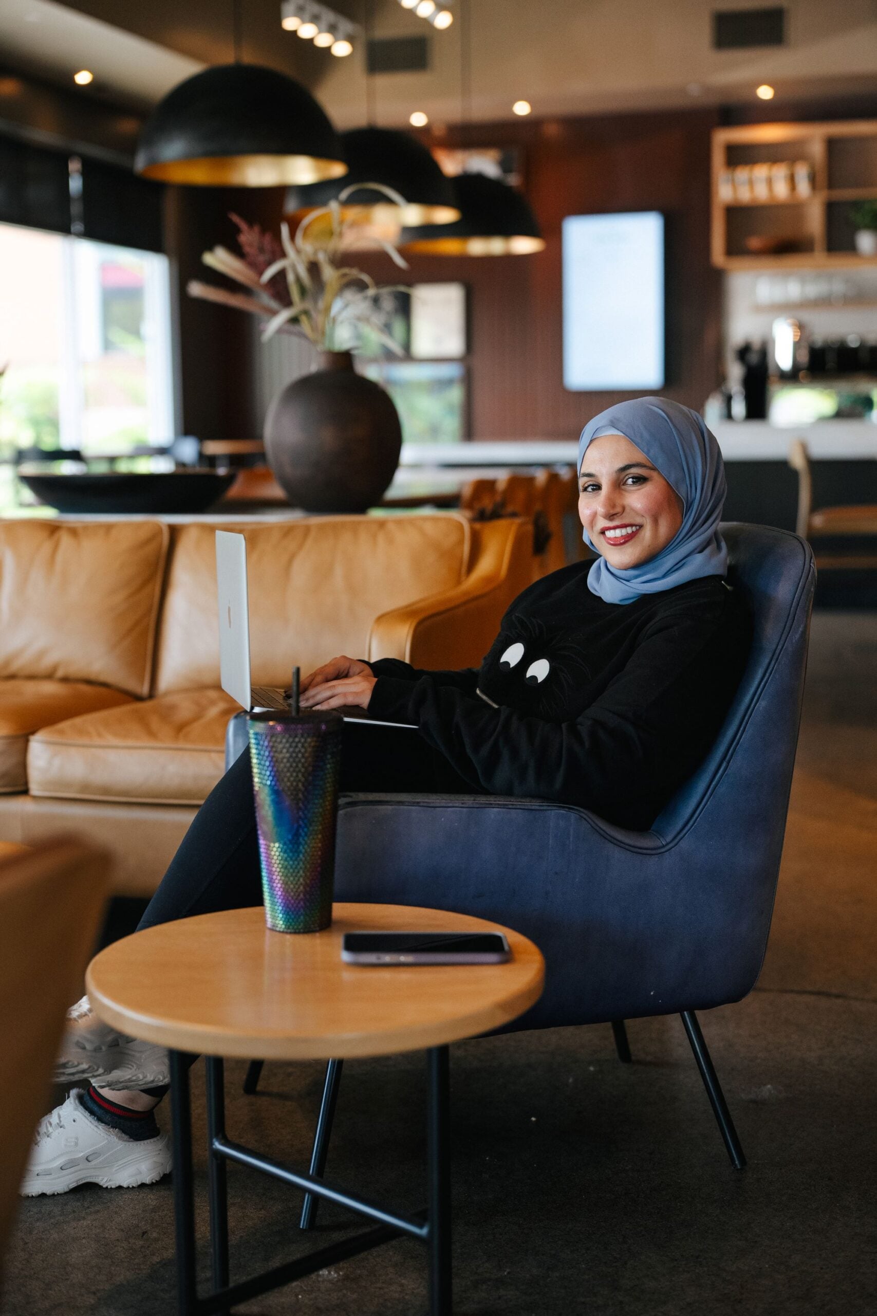 A woman in a blue hijab sits in a cozy café, smiling while using a laptop. A colorful cup and a phone rest on a small table nearby.