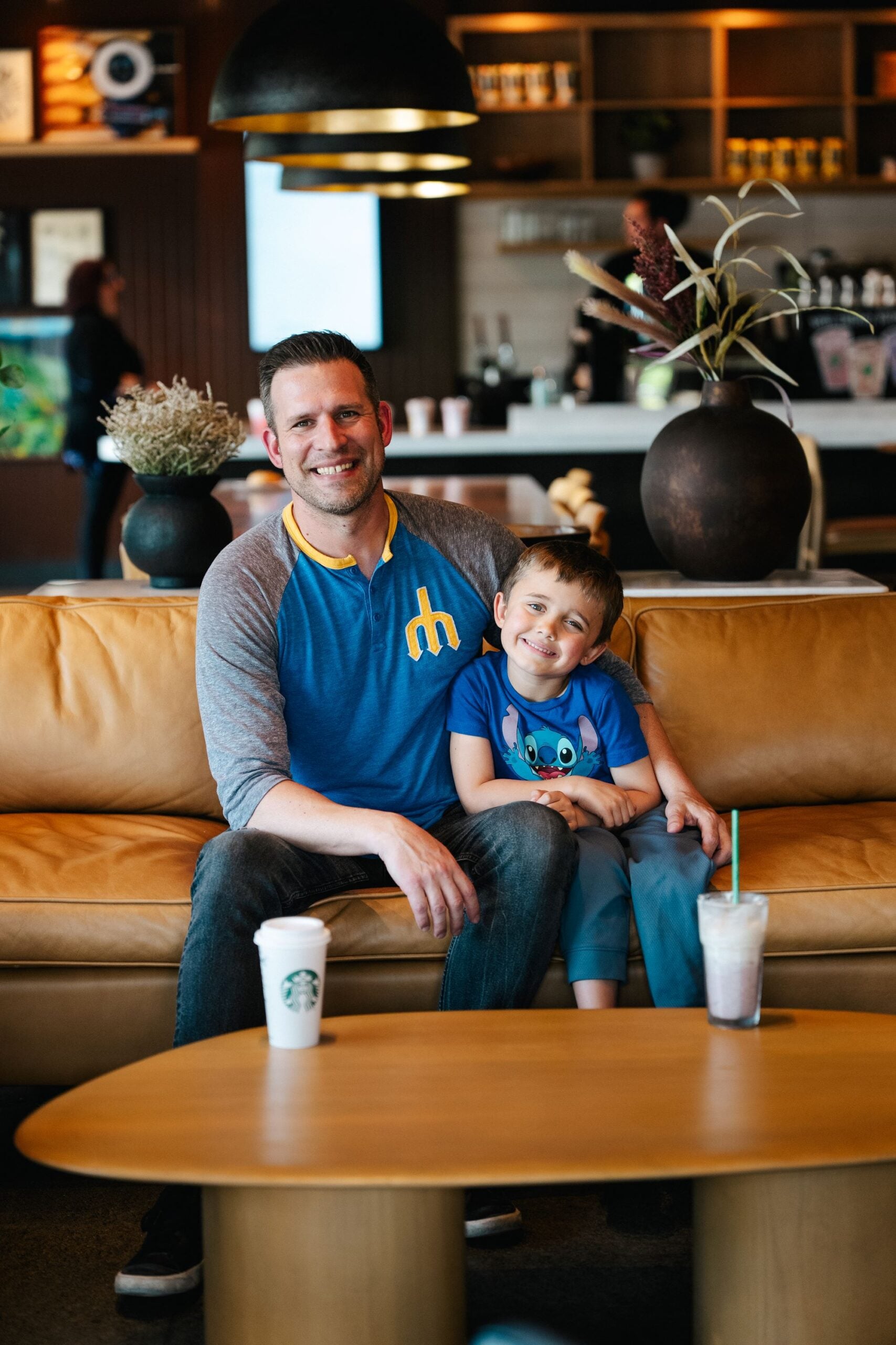 A man and a boy sit together on a brown leather couch, smiling. The man wears a blue shirt with gray sleeves, while the boy is in a blue shirt featuring a cartoon character. Two drinks are on a round wooden table in front of them. The background shows a coffee shop setting.