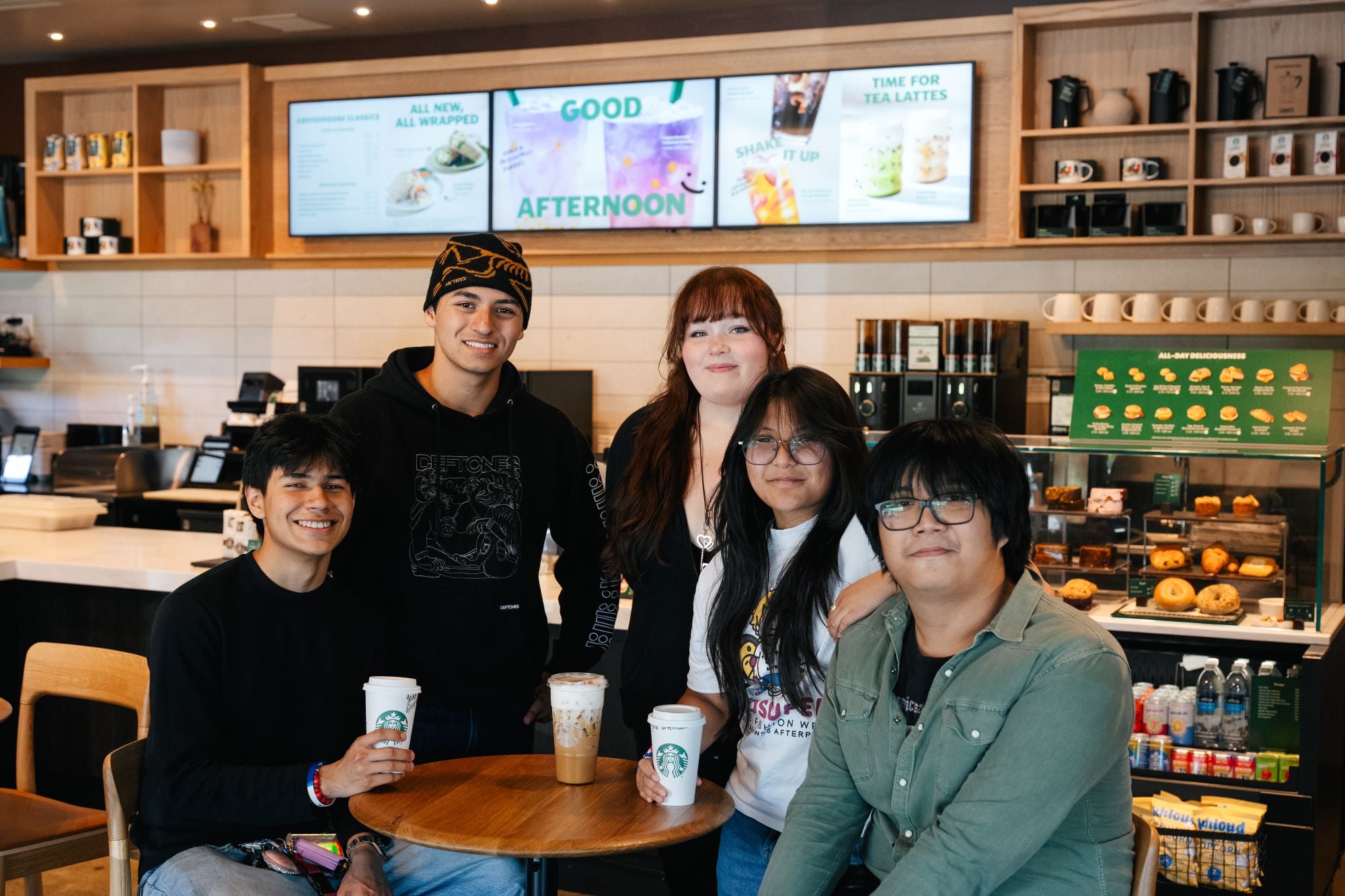 A group of five young adults poses together at a Starbucks, holding drinks. The café features a modern design with menu boards and a display of pastries in the background.