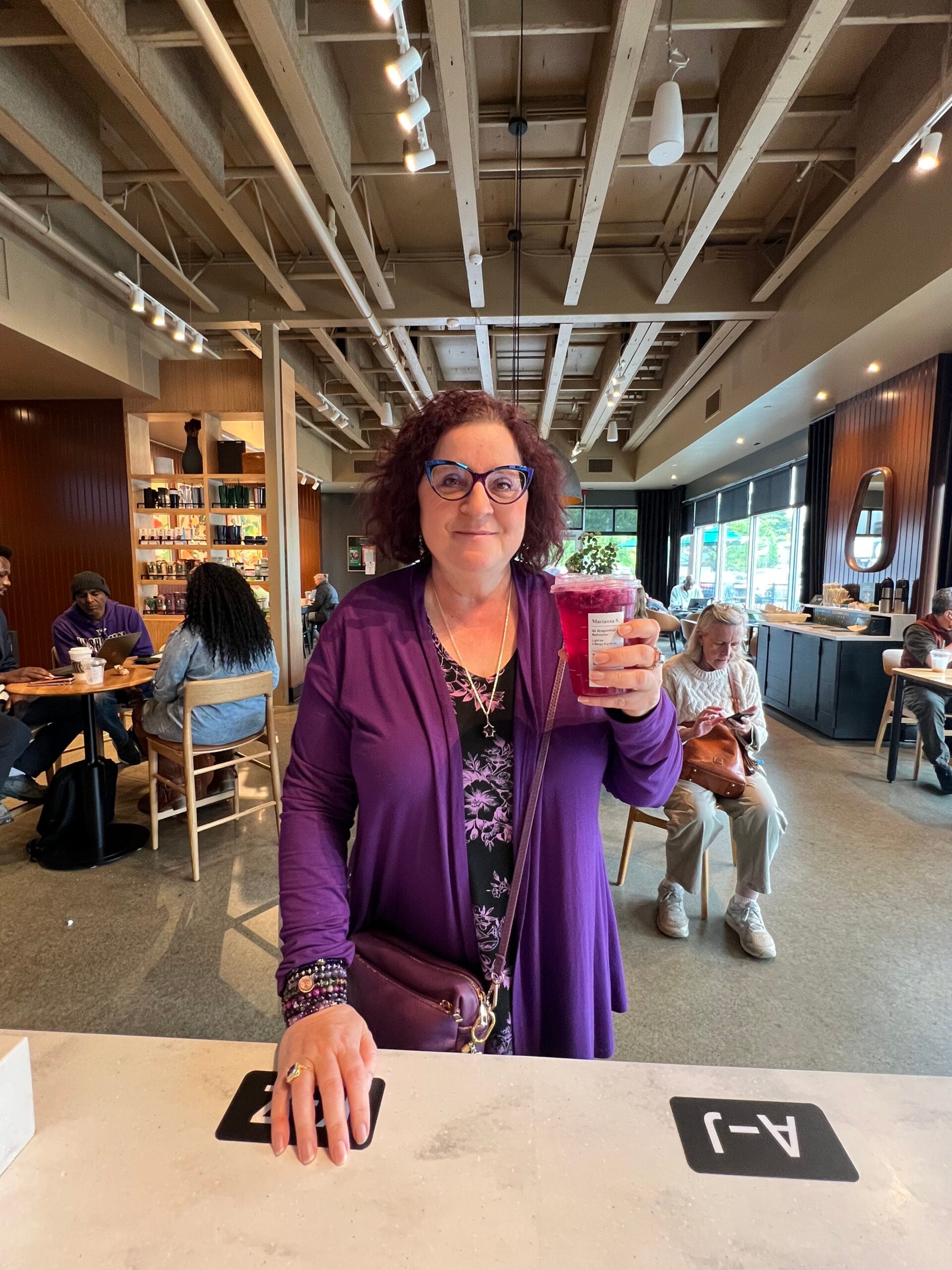 A woman with curly hair, wearing glasses and a purple outfit, holds a drink at a café. The interior features wooden beams and seating.