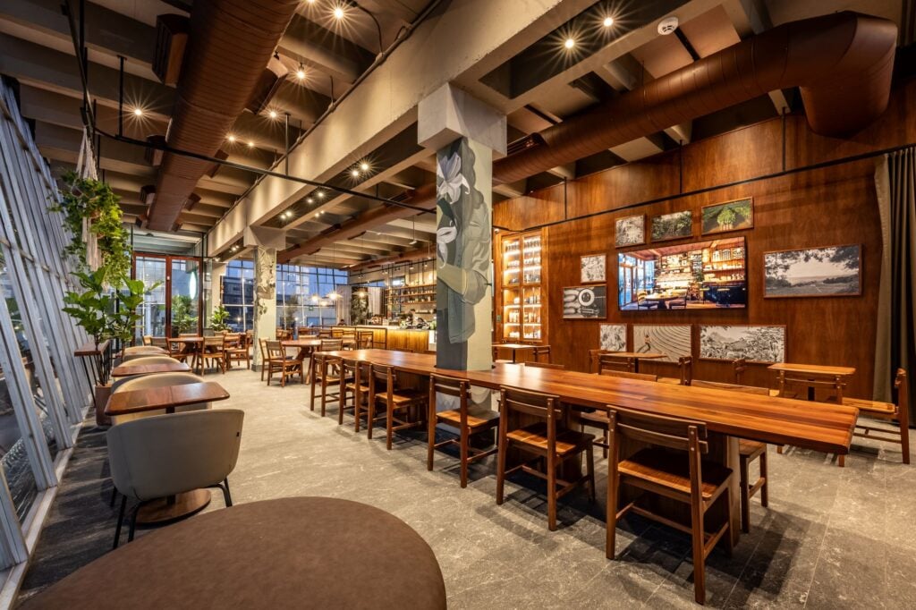 A modern café interior featuring wooden tables and chairs, large windows, and a bar area with shelves. Green plants add a touch of nature.