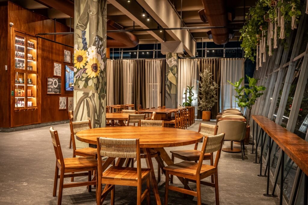 A modern café interior featuring wooden tables and chairs, decorative floral murals, and large windows with soft curtains.
