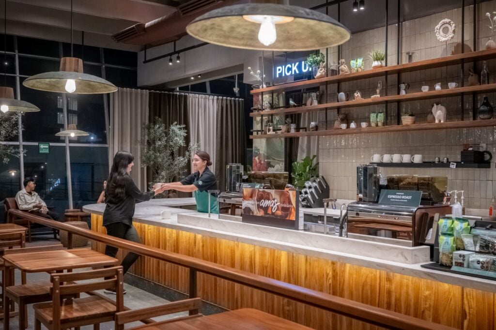 A modern café interior features a marble counter where a barista hands a drink to a customer. Shelves display decorative items and plants. Soft lighting and wooden furniture create a cozy atmosphere.