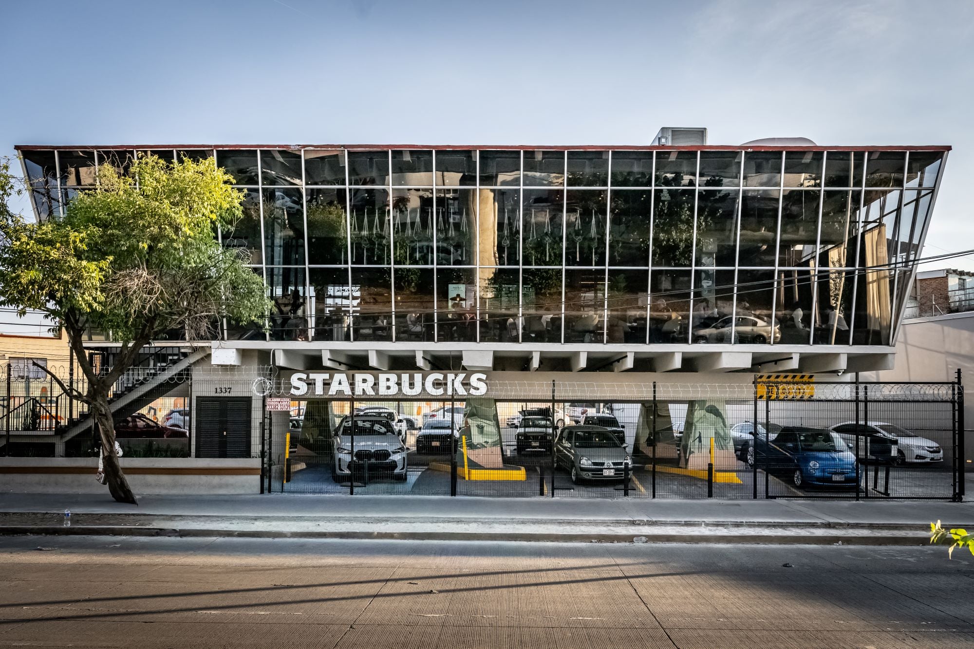 A modern Starbucks building features a glass facade with greenery visible inside. The lower level has parking spaces for several cars.