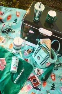 A vibrant picnic setup features a teal blanket adorned with colorful Starbucks-themed stickers. A green cooler sits atop, accompanied by various drinkware, including a tumbler and a mug. Nearby, a small bag holds a map, and scattered around are playing cards and instant photos, creating a lively outdoor atmosphere.