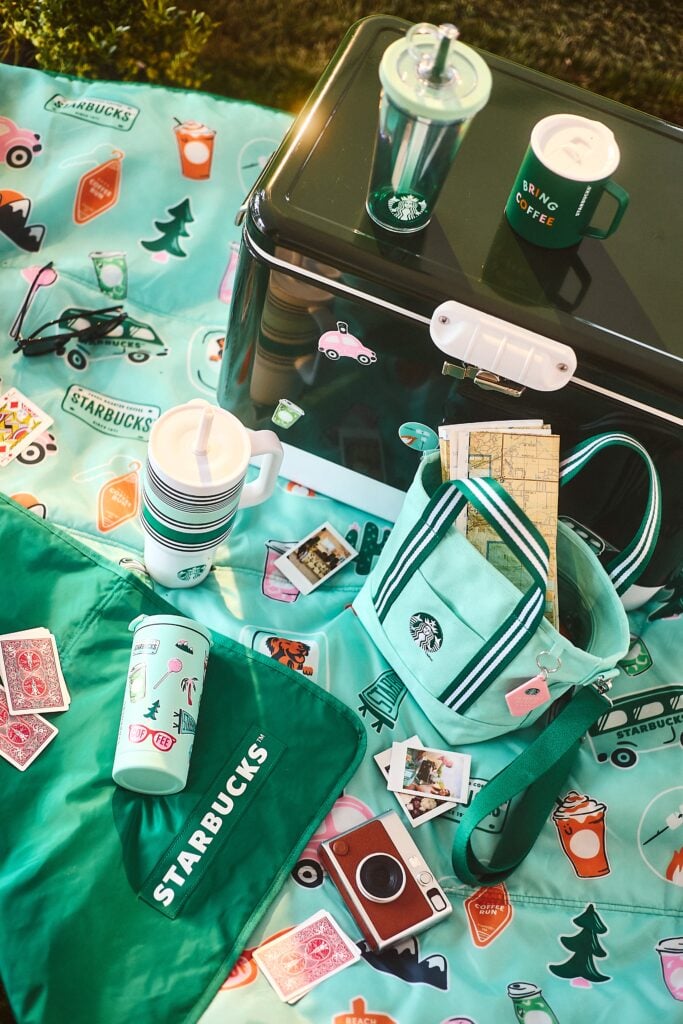 A vibrant picnic setup features a teal blanket adorned with colorful Starbucks-themed stickers. A green cooler sits atop, accompanied by various drinkware, including a tumbler and a mug. Nearby, a small bag holds a map, and scattered around are playing cards and instant photos, creating a lively outdoor atmosphere.