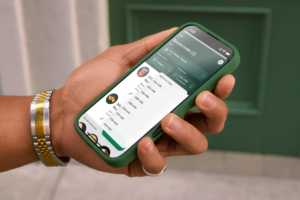 A hand holds a smartphone with a green case displaying a food order review screen. The screen shows items, prices, and estimated pickup times. A wristwatch with a silver and gold band is visible. The background features a green door.