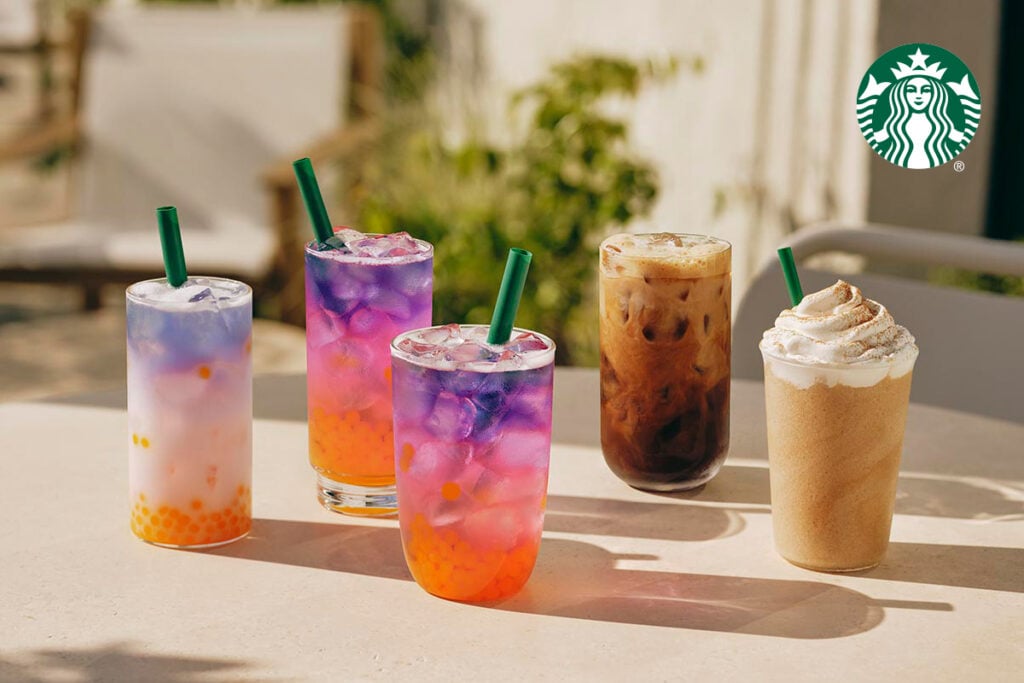 Five colorful drinks are displayed on a table, featuring vibrant layers and toppings. Three have green straws, while two are iced coffee variations.