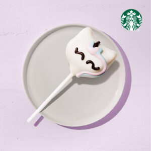 A cake pop shaped like a ghost, coated in white chocolate with colorful icing details, sits on a light gray plate against a pastel background.