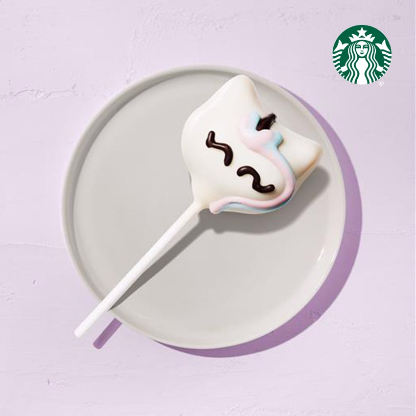 A cake pop shaped like a ghost, coated in white chocolate with colorful icing details, sits on a light gray plate against a pastel background.