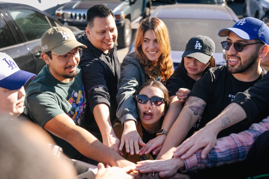 A group of people is gathered with their hands stacked together in a circle, expressing excitement and camaraderie. They are smiling and engaged, with a mix of casual clothing and accessories, including hats and sunglasses. In the background, vehicles are parked, suggesting an outdoor setting.