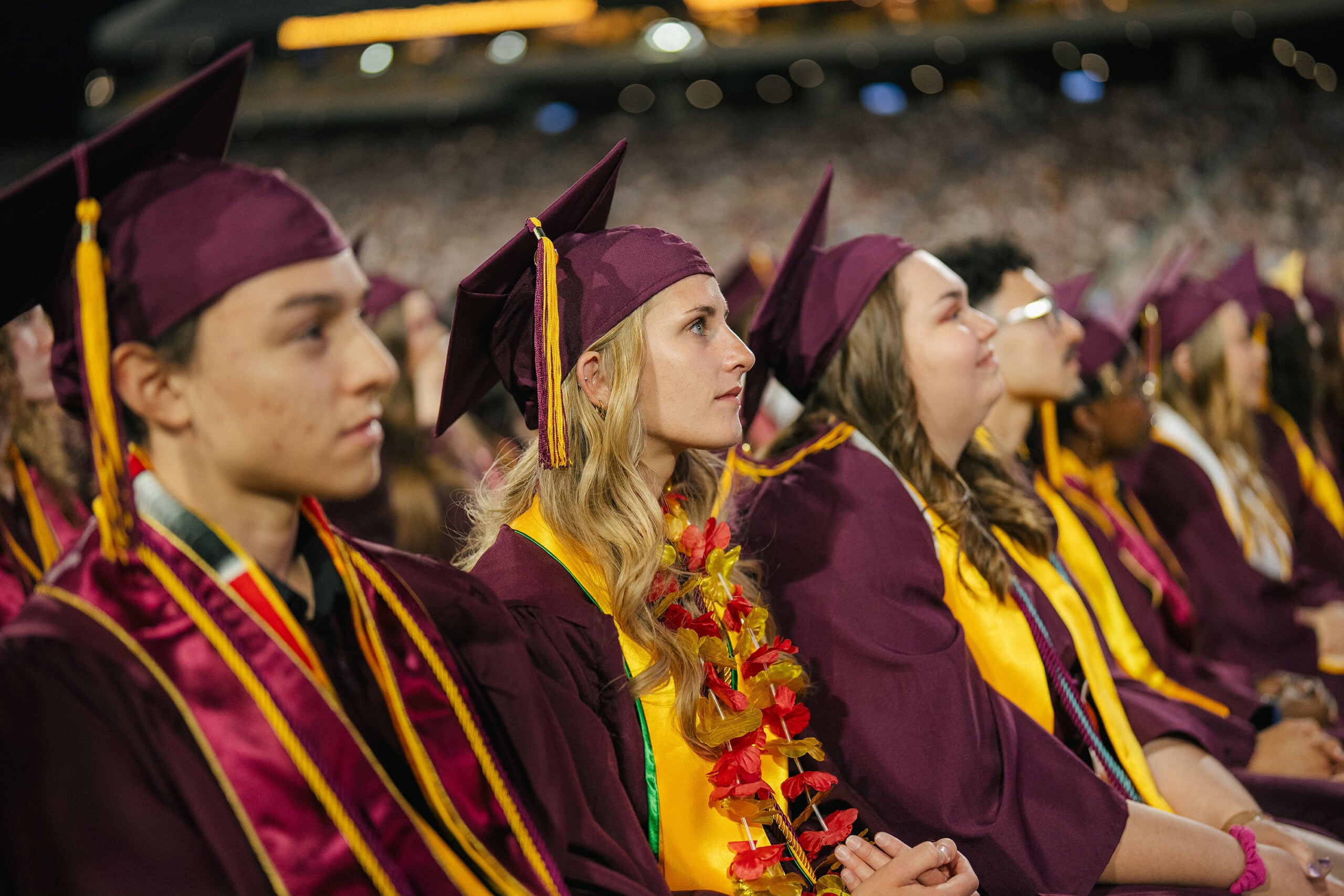 Graduates in maroon caps and gowns sit in a stadium, focused on the ceremony. One wears a floral lei, adding a colorful touch.