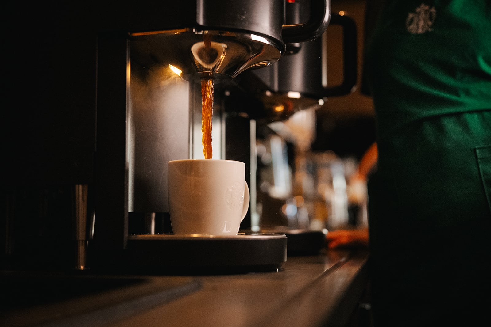 Coffee is being brewed and poured into a white cup on a counter, with a barista in a green apron in the background.