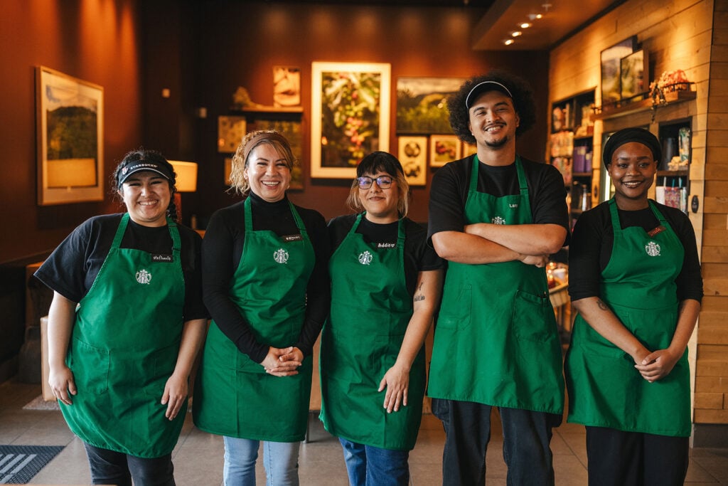 Five baristas stand together in a coffee shop, all wearing green aprons. The background features warm lighting and framed artwork.