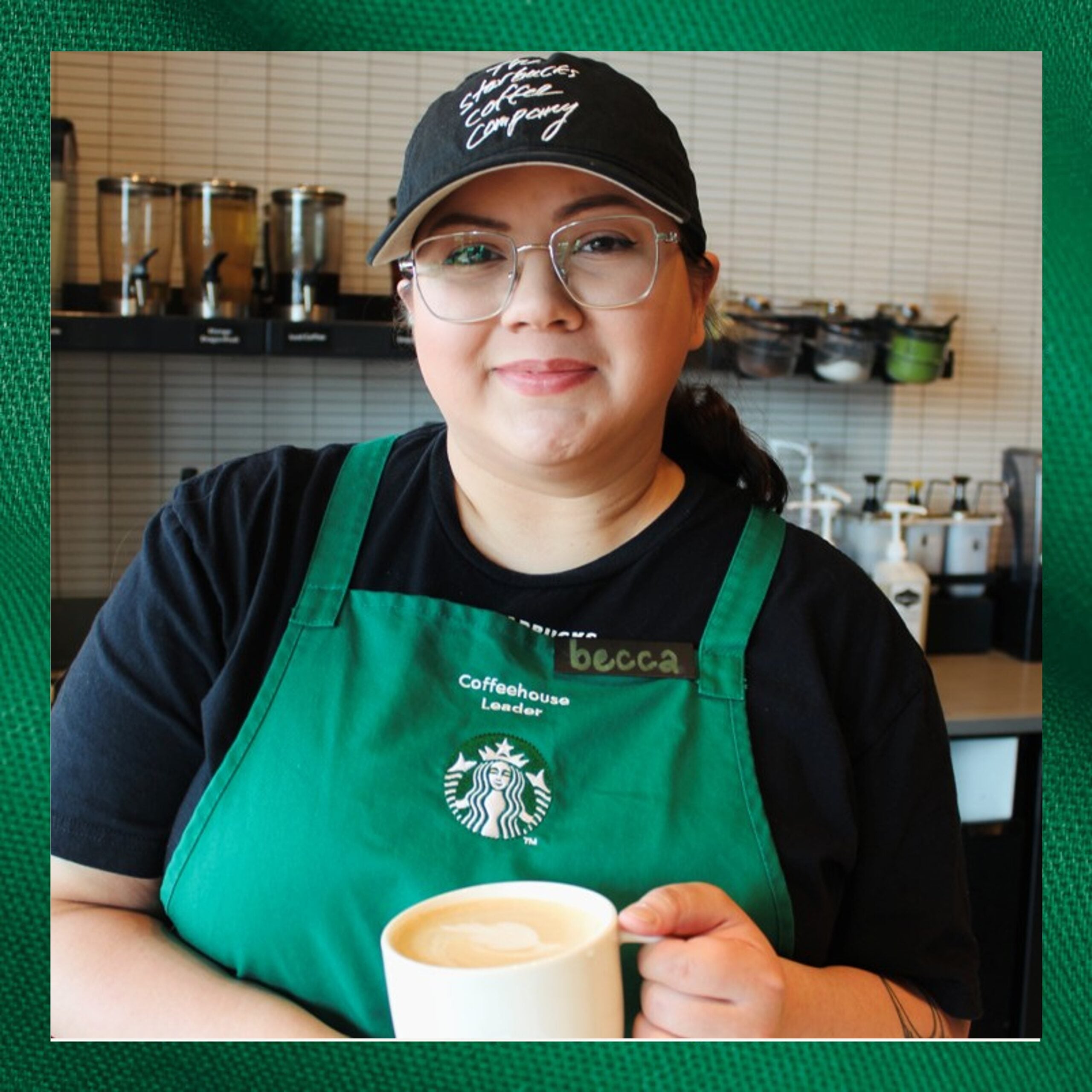 A barista wearing a green apron and a black cap smiles while holding a white coffee cup. The background features coffee equipment.