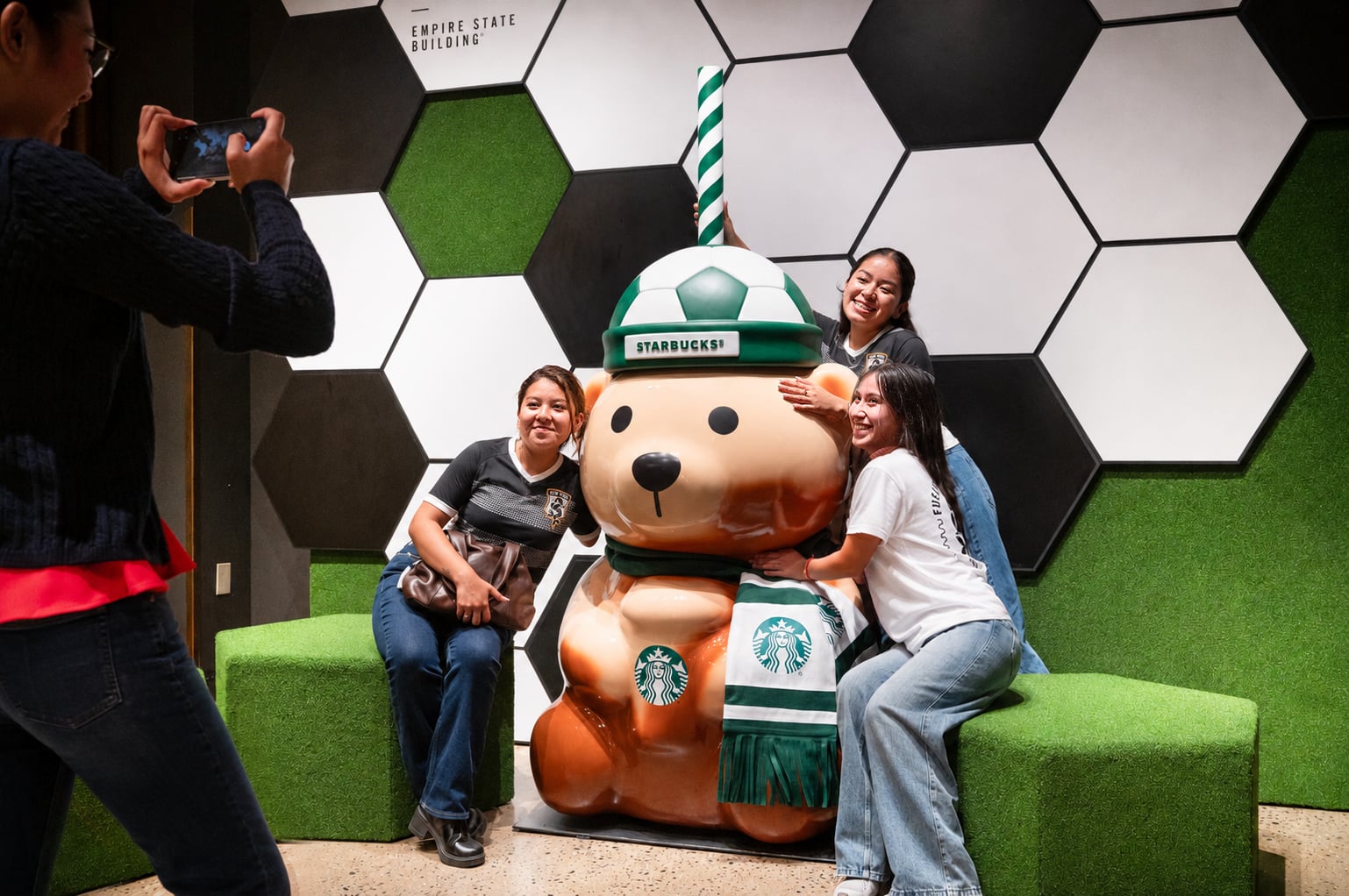 Four people pose with a large, bear-shaped statue wearing a soccer-themed Starbucks hat and scarf. The background features hexagonal patterns and green accents.