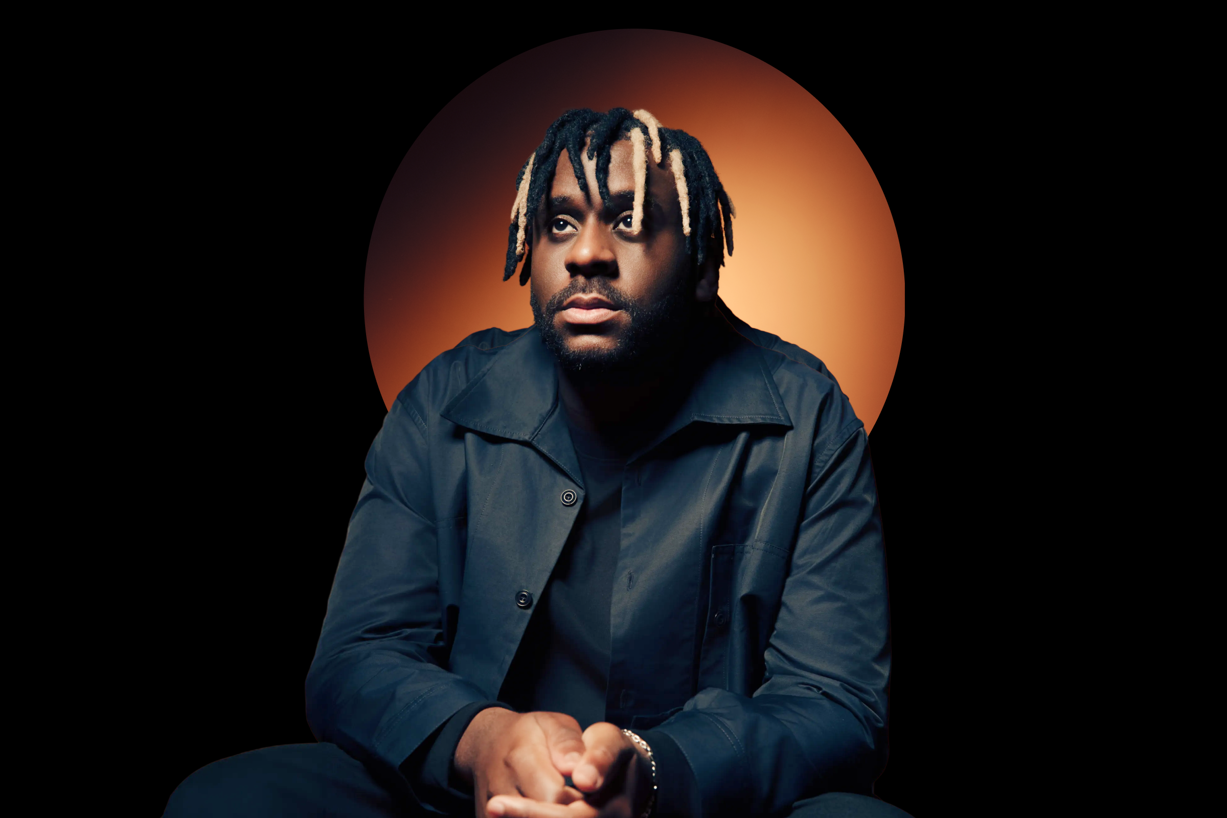 A person with textured hair styled in loose strands sits in front of a warm, circular background. They wear a dark jacket and appear contemplative.