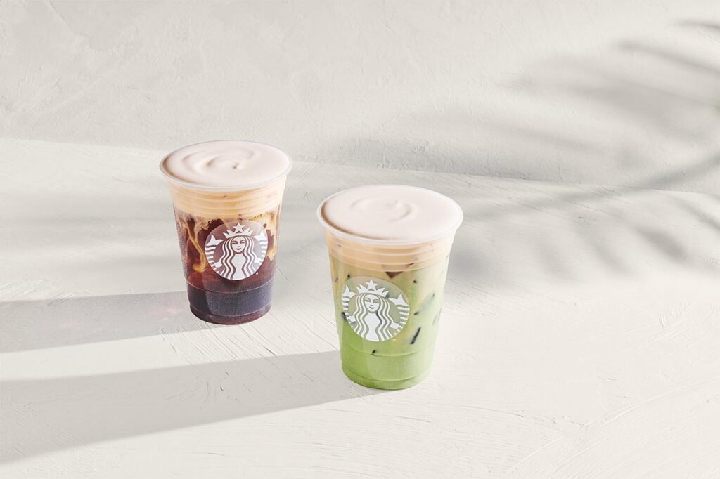 Two iced beverages are displayed on a light surface. One is dark brown with a creamy top, while the other is green with a similar creamy layer. Both cups feature a logo.