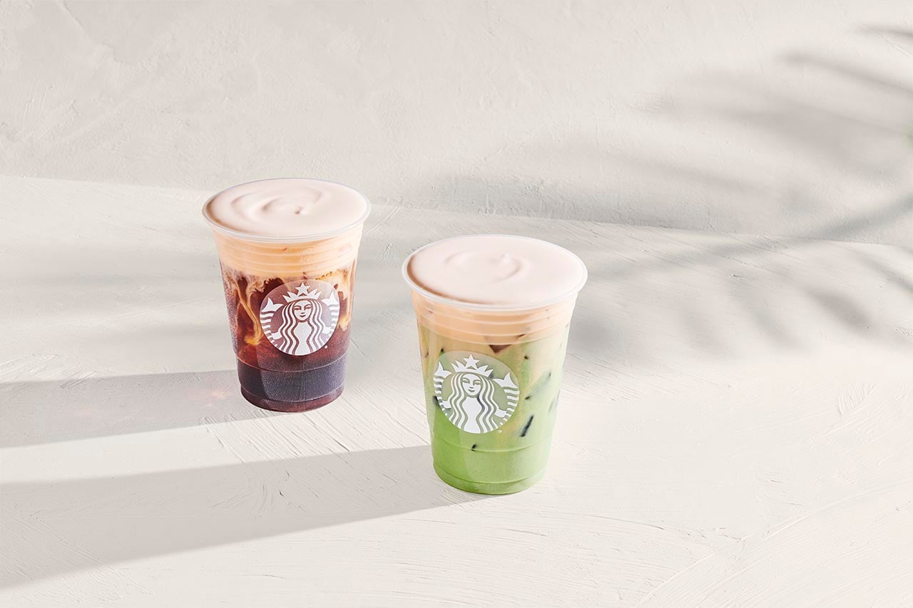 Two iced beverages are displayed on a light surface. One is dark brown with a creamy top, while the other is green with a similar creamy layer. Both cups feature a logo.