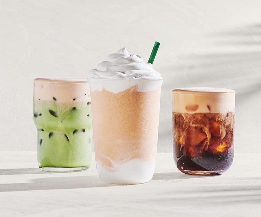 Three beverages are displayed: a green iced drink with a pink top, a creamy drink topped with whipped cream and a straw, and a dark iced coffee with a frothy layer.