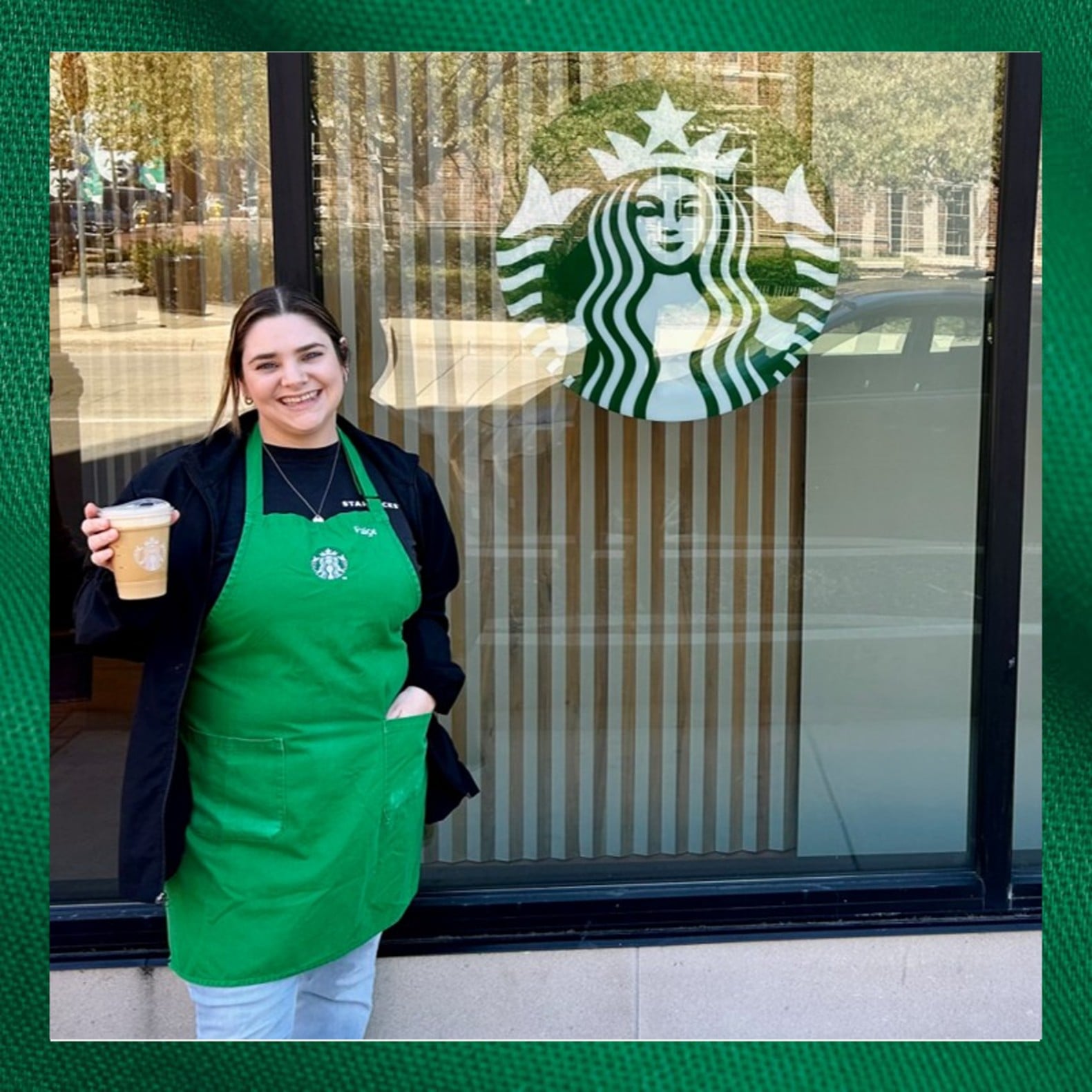A barista in a green apron stands outside a Starbucks, holding a coffee cup. The Starbucks logo is visible on the window behind her.