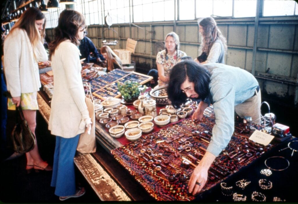 A market scene featuring a vendor arranging colorful jewelry and pottery on a table. Several shoppers browse the items.