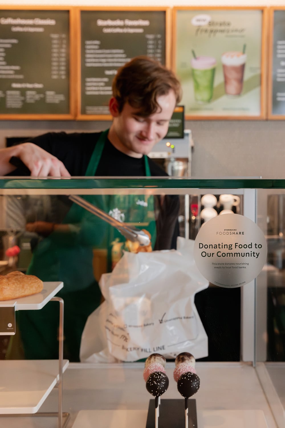 A barista in a green apron is serving customers at a café. In the display case, there are pastries and cake pops. A sign highlights a food donation initiative.