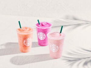 Three colorful smoothies in clear cups with green straws are displayed. The flavors appear to be pink, orange, and light pink, each featuring a logo on the front. The background is light and textured, suggesting a bright, summery vibe.