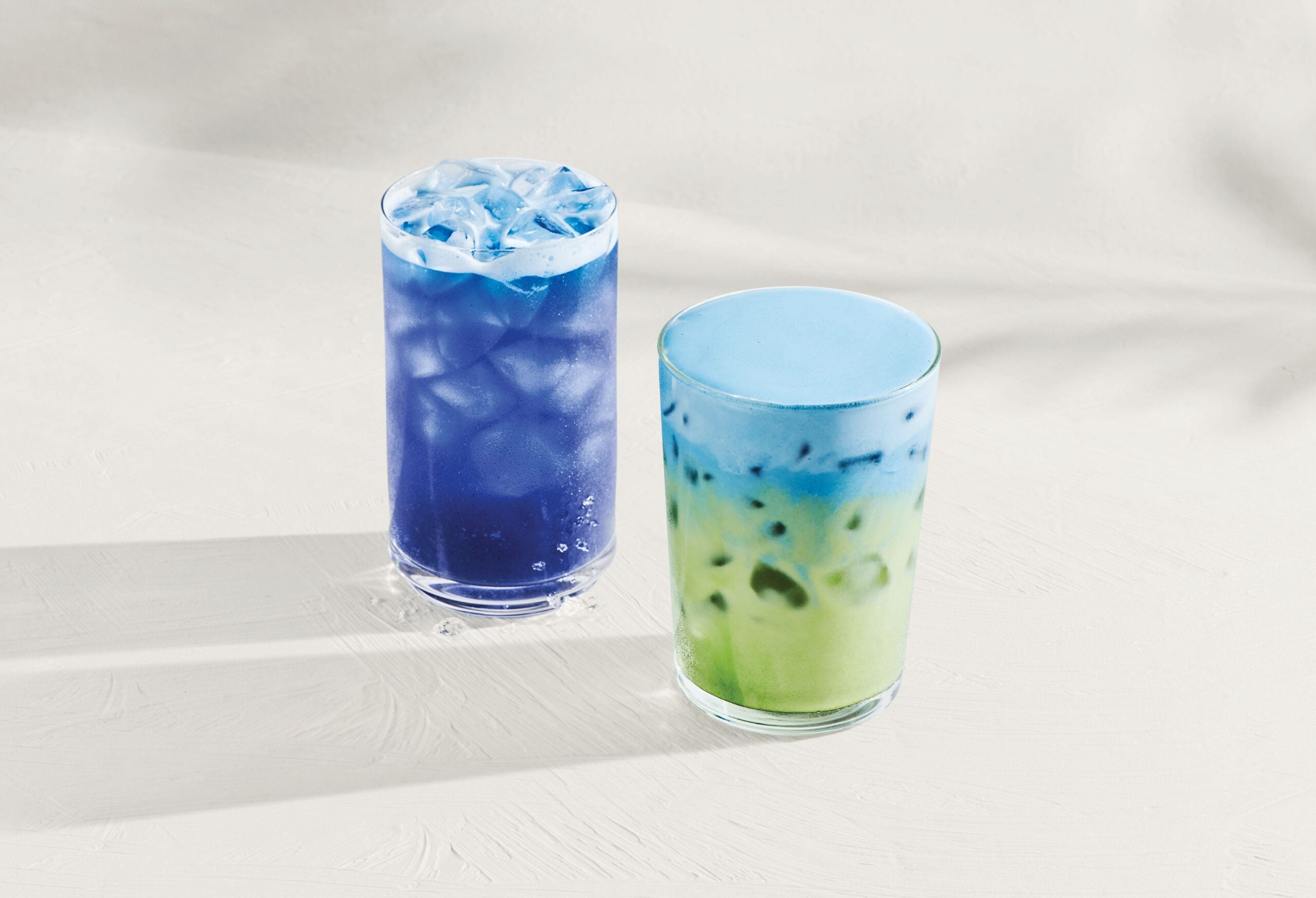Two colorful drinks are displayed on a light surface. The left drink is a vibrant blue with ice, while the right drink features a gradient of blue to green, also with ice.