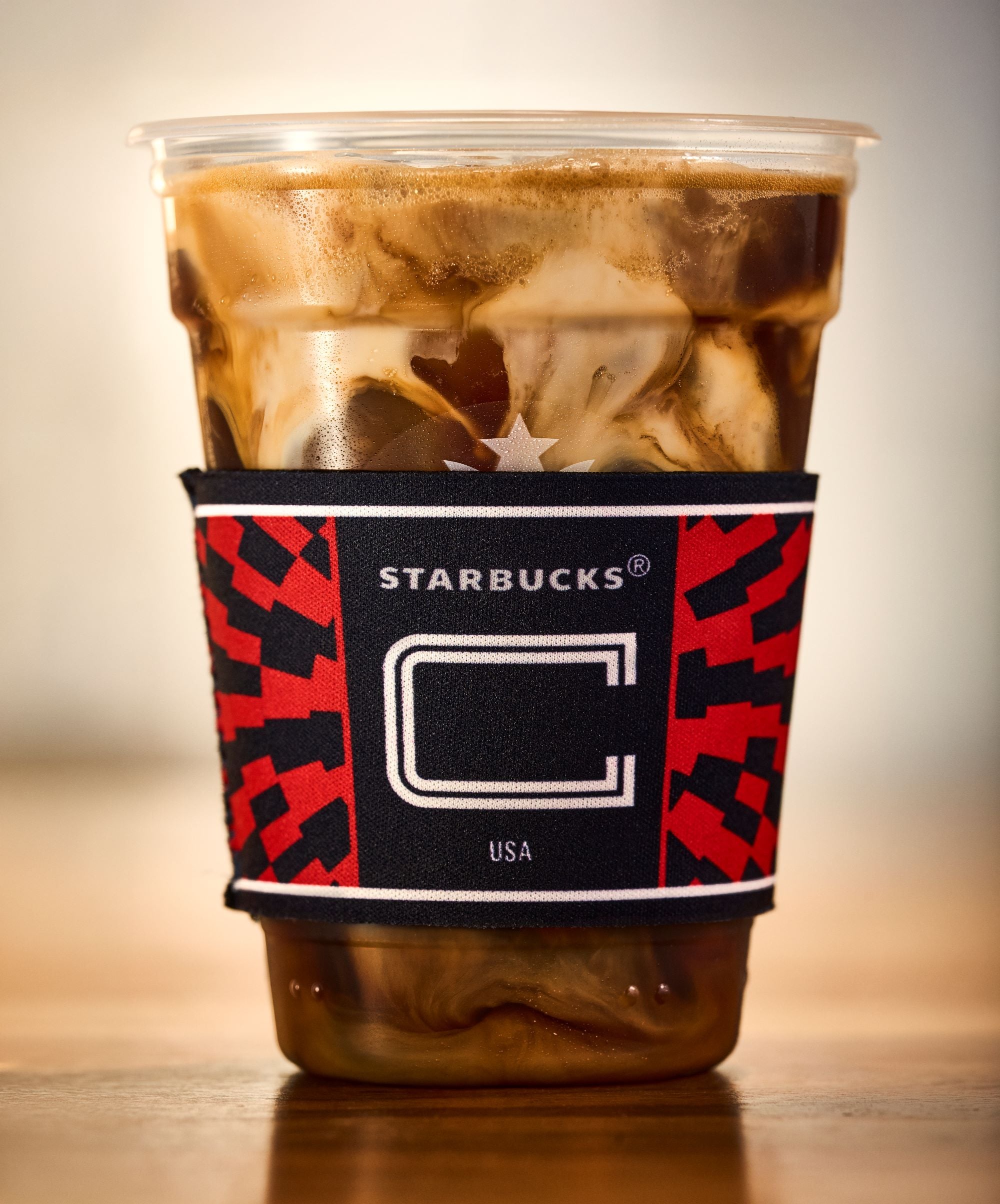 A clear cup filled with iced coffee, featuring swirling cream, is wrapped in a black and red Starbucks sleeve.