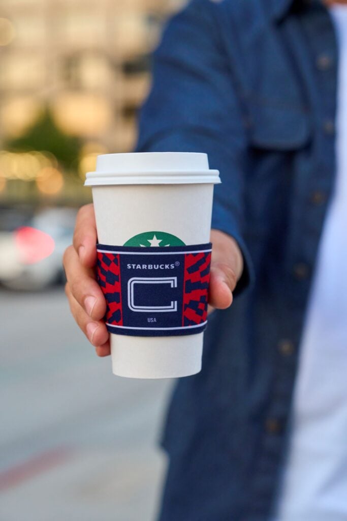 A person is holding a Starbucks coffee cup with a colorful sleeve featuring a design in red and blue. The background is blurred, suggesting an outdoor setting.