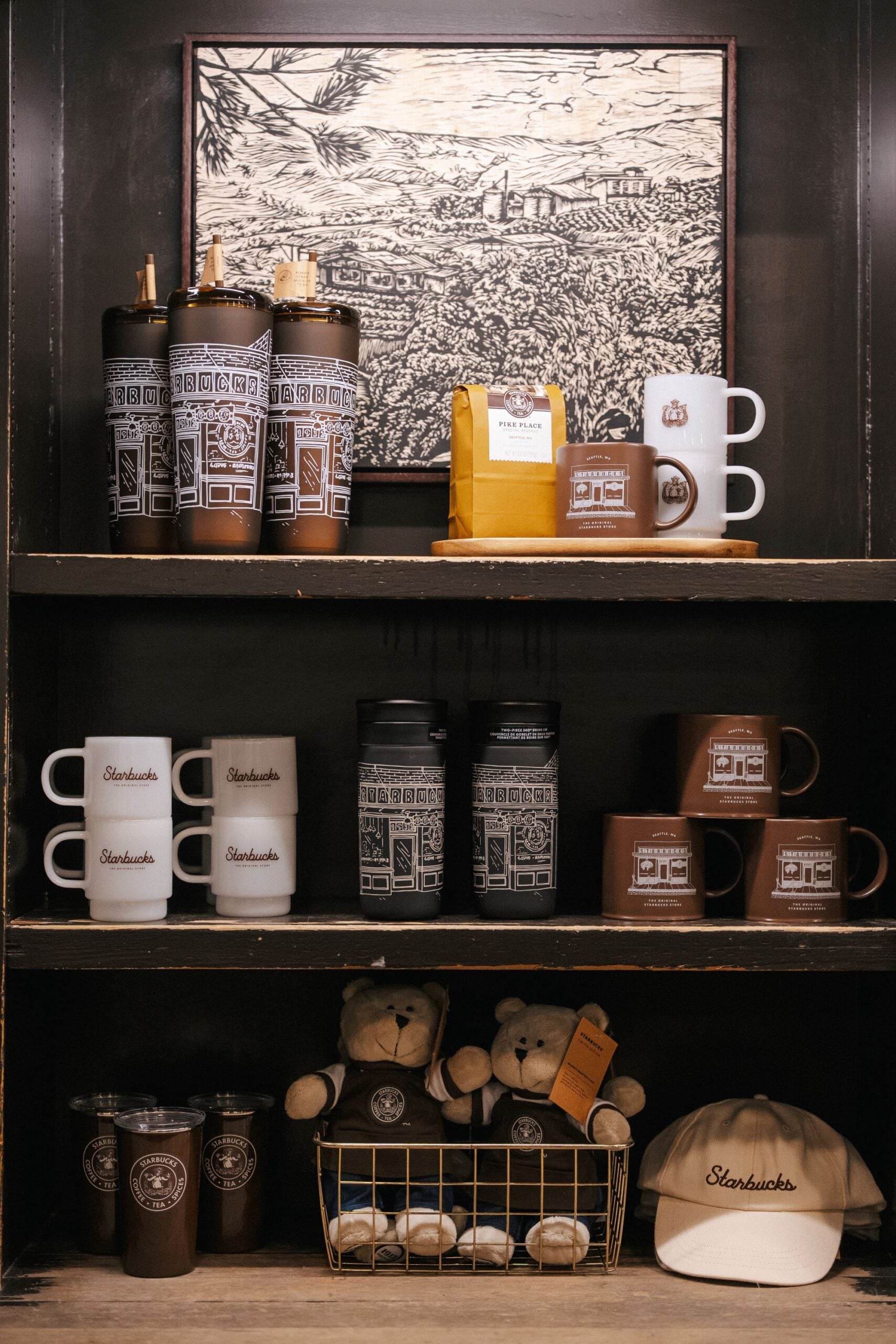 A display shelf features various Starbucks merchandise, including travel mugs, coffee mugs, a bag of coffee, and plush teddy bears, alongside a baseball cap. The items are arranged neatly, showcasing a mix of colors and designs.