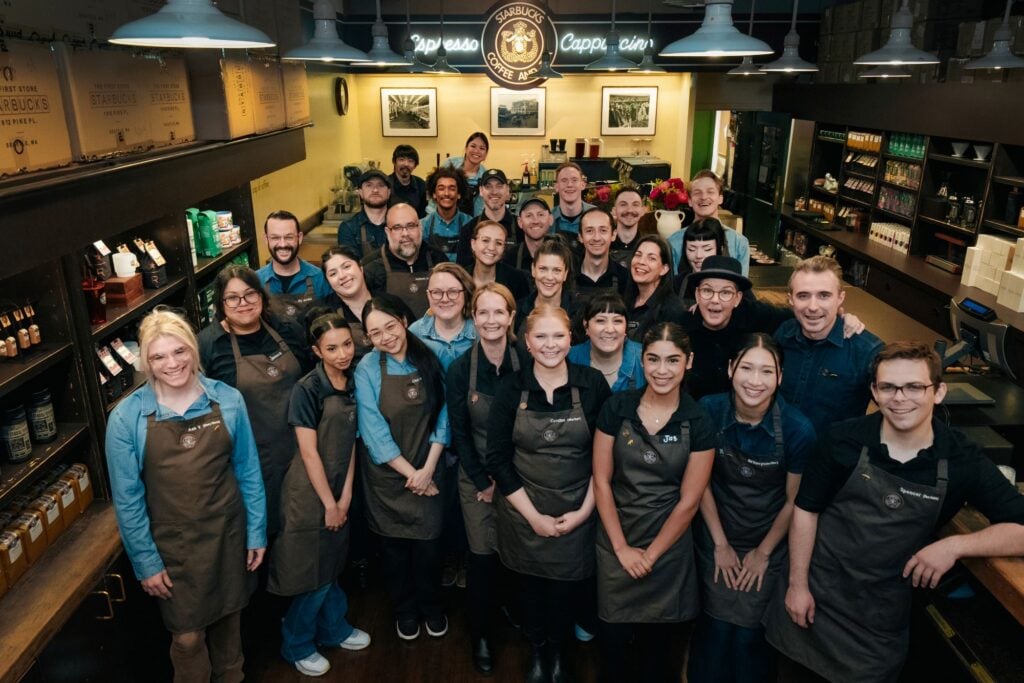 A large group of baristas and staff members pose together in a coffee shop, wearing brown aprons. The shop features shelves of coffee products and a sign that reads "Espresso Cappuccino.