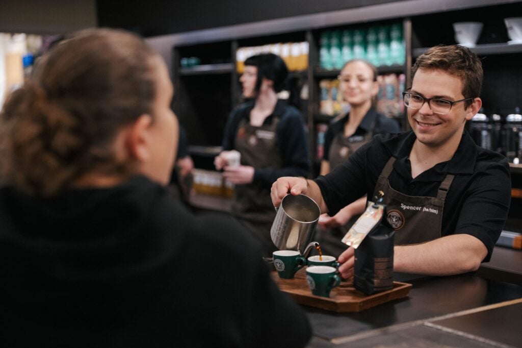 A barista is pouring coffee into two small green cups on a wooden tray. Several other staff members are visible in the background.