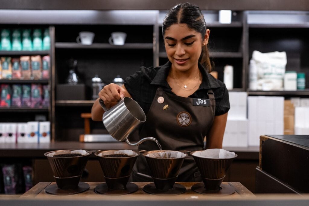 A barista pours hot water from a metal kettle into multiple coffee drippers filled with ground coffee, smiling as she works.