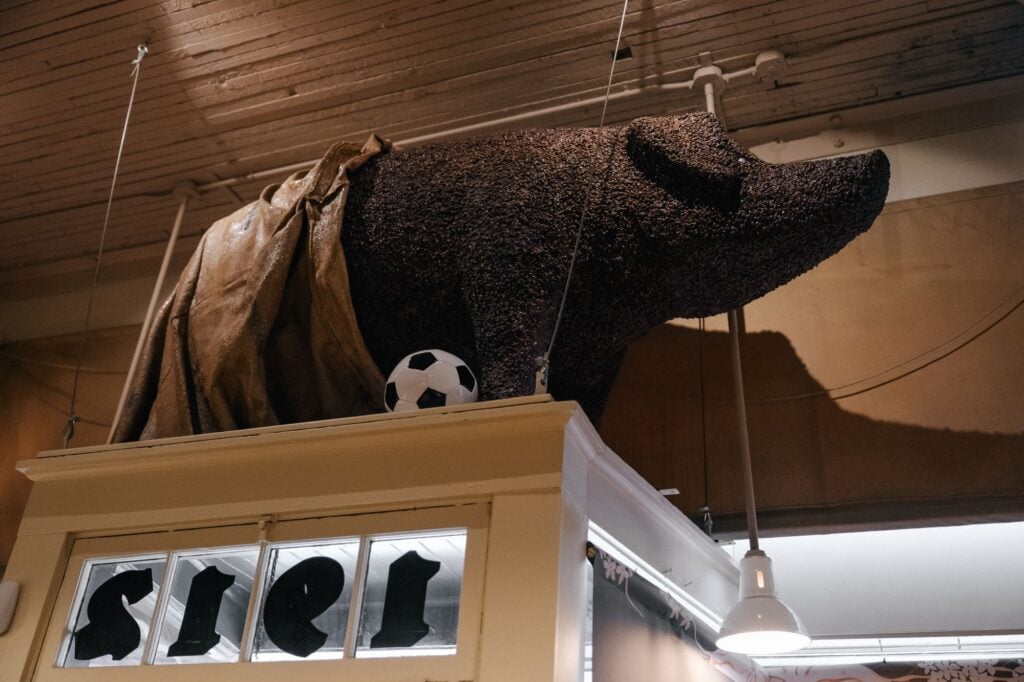 A large, textured sculpture of a pig draped in fabric is displayed above a storefront. A small soccer ball rests nearby.