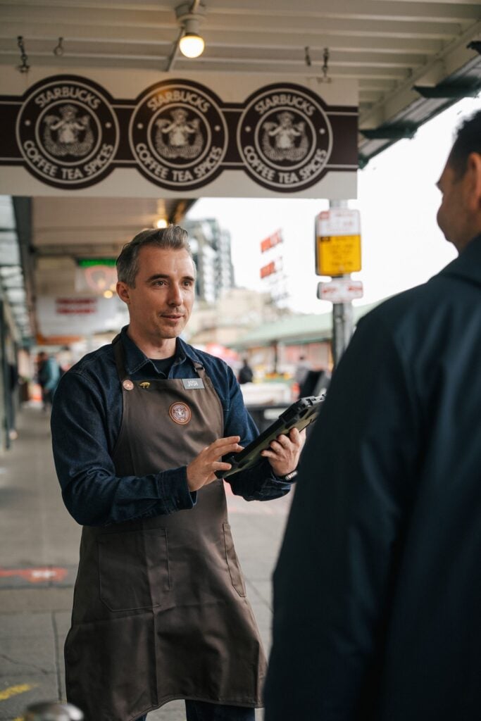 A barista in a brown apron holds a tablet while engaging with a customer outside a Starbucks, with signage overhead.