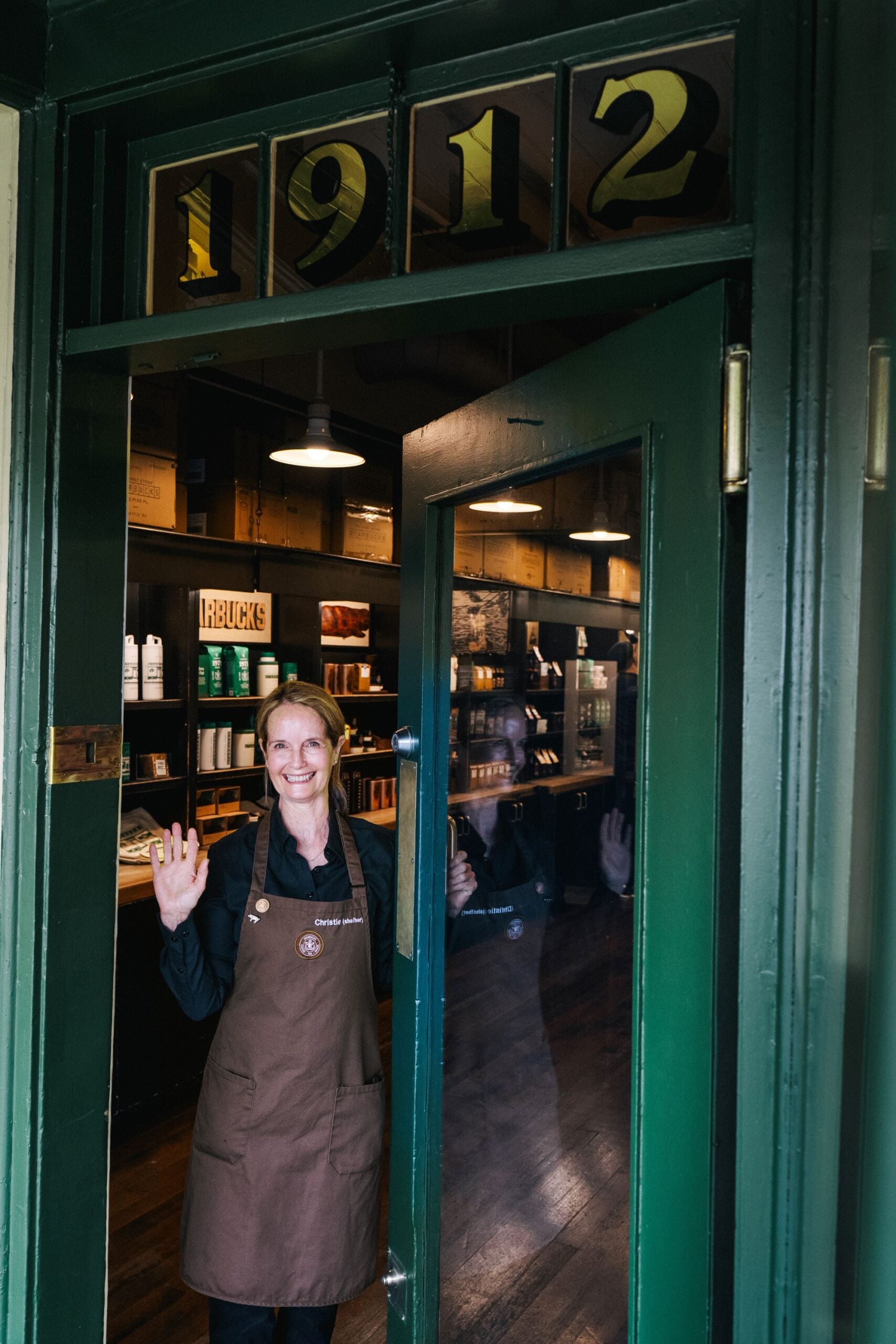 A woman in a brown apron stands at an open green door, smiling and waving. The background features shelves with products and a sign reading "1912.