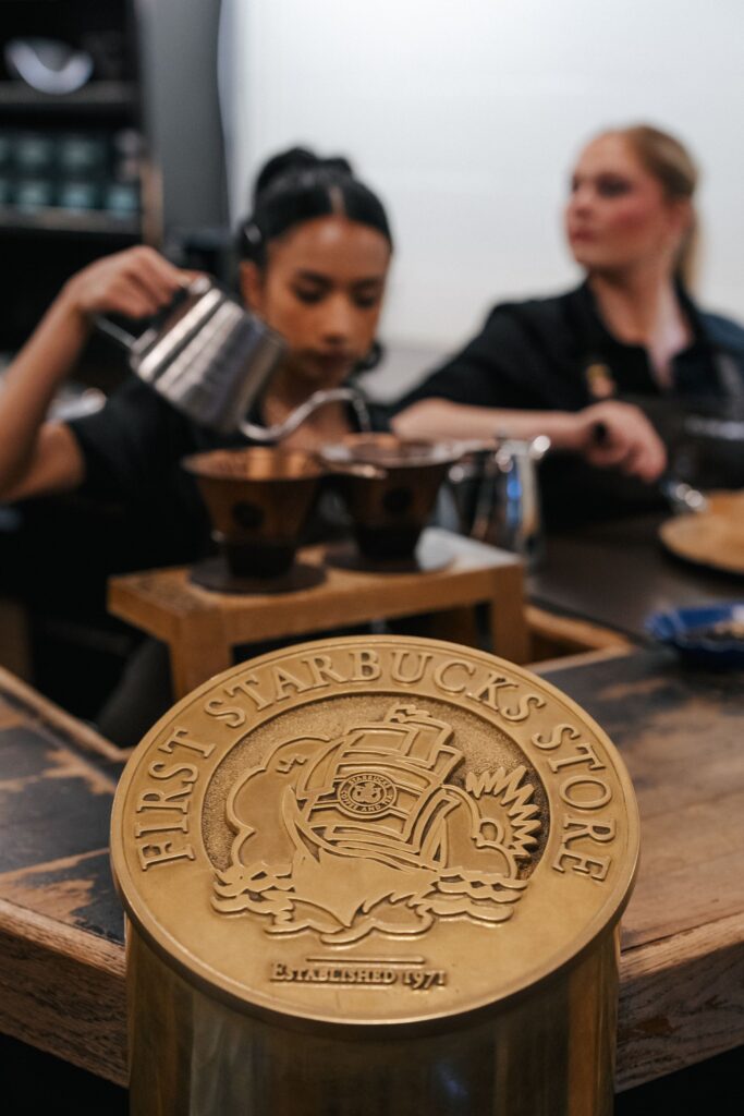 A brass plaque reading "FIRST STARBUCKS STORE ESTABLISHED 1971" is in focus, with baristas brewing coffee in the background.