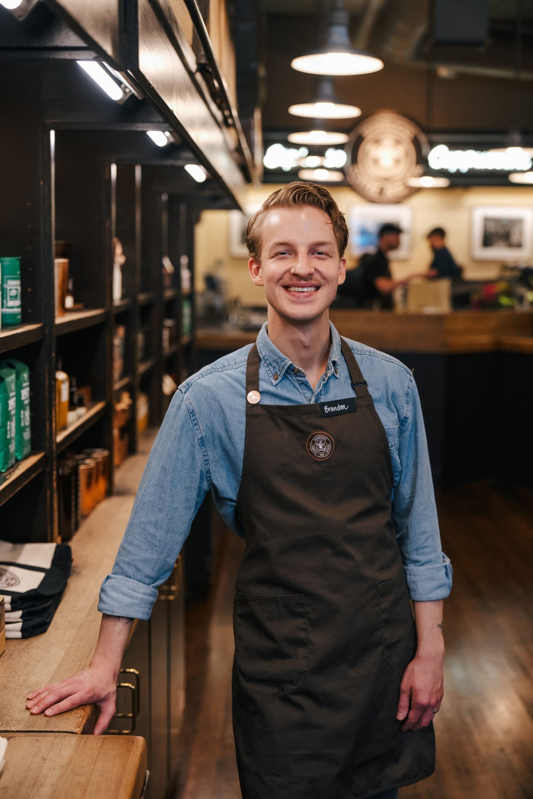 A smiling barista stands in a coffee shop, wearing a denim shirt and a dark apron. Shelves filled with coffee products are in the background.