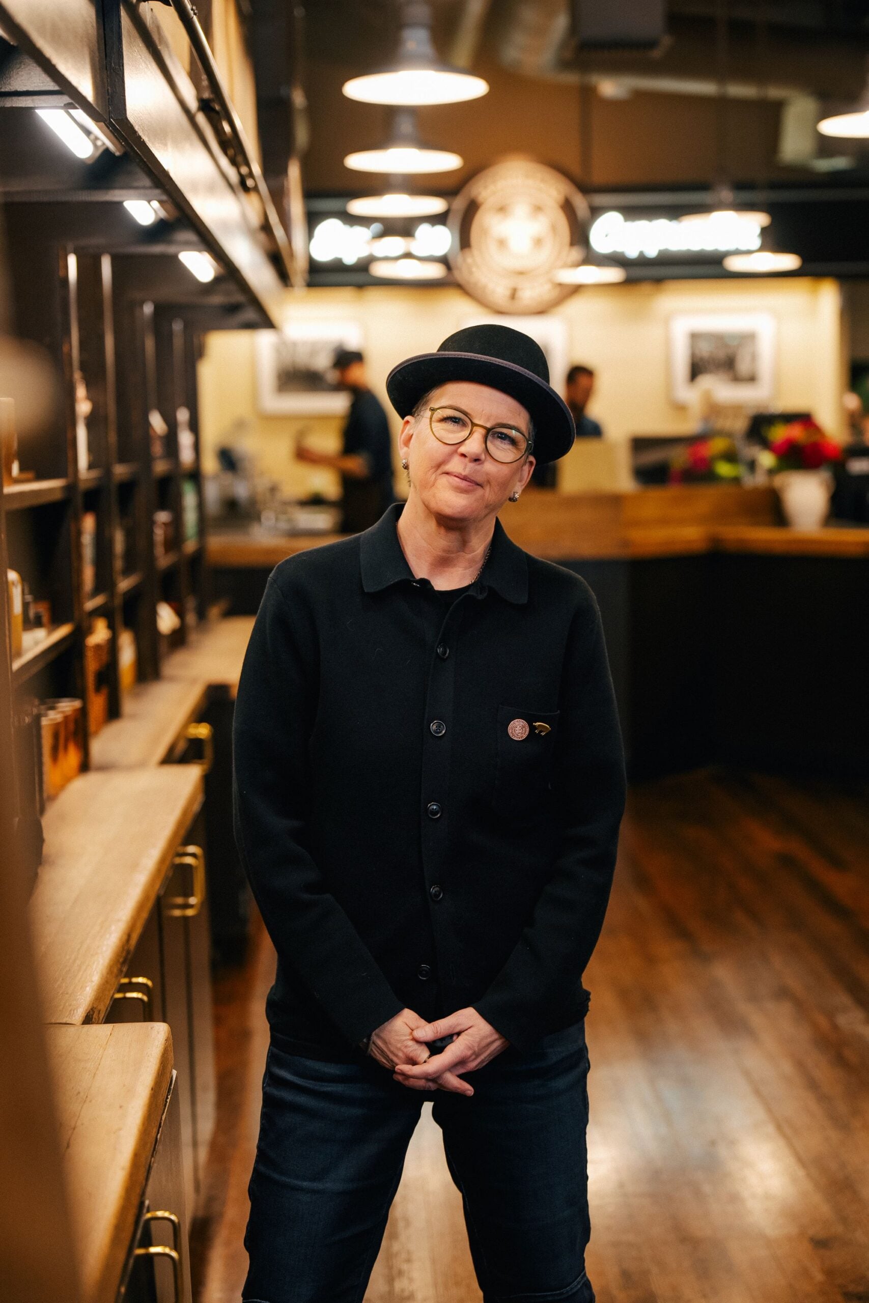 A person stands in a coffee shop, wearing a black cardigan and a black hat. The background features wooden shelves and a counter with staff.
