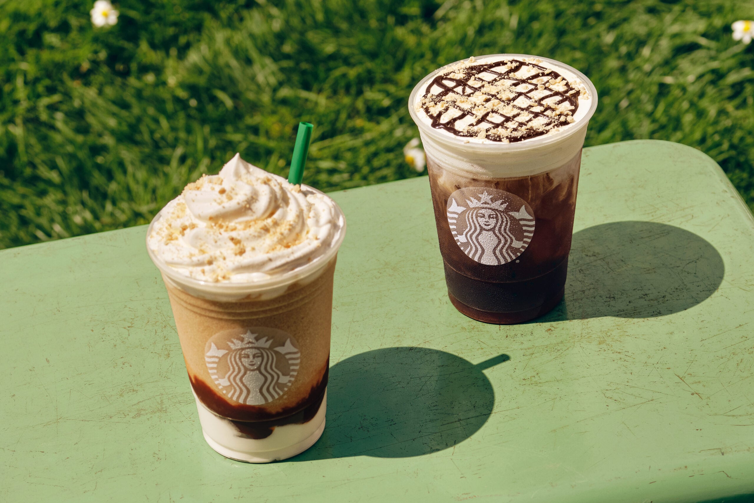 Two Starbucks beverages sit on a green table. One is a blended drink topped with whipped cream and graham cracker crumbs, while the other is a dark iced drink with whipped cream and chocolate drizzle. The background features grass.