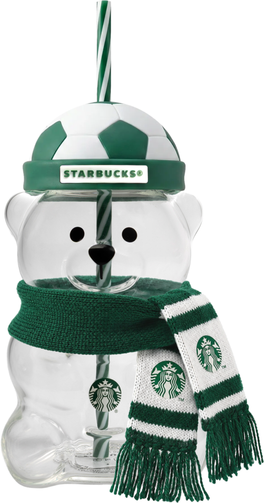 A clear bear-shaped cup features a soccer ball lid and a green and white striped straw. It wears a knitted scarf with fringe.