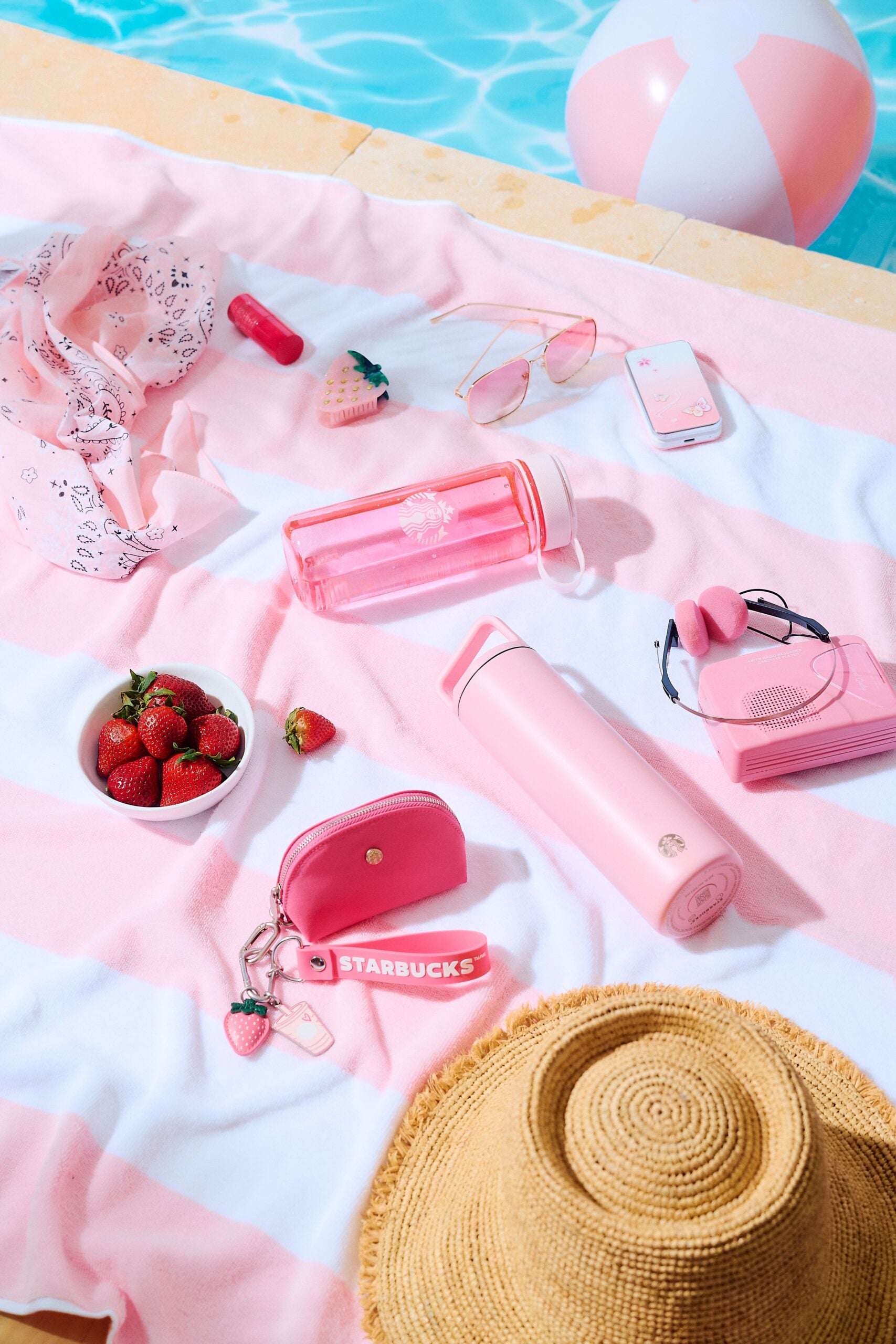 A pink and white striped towel is spread out, featuring a variety of pink accessories: a water bottle, a thermos, a small wallet, sunglasses, a phone, headphones, and a beach ball. A bowl of strawberries and a straw hat complete the summer vibe.