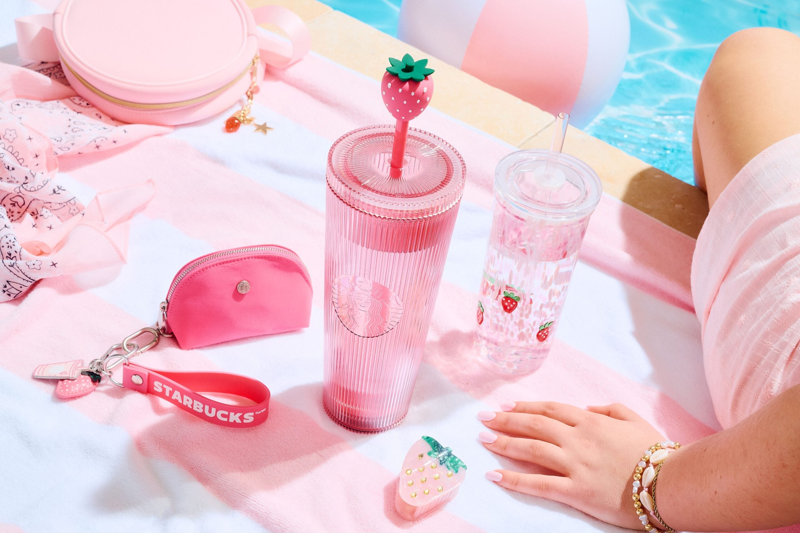 A pink striped towel features a tall, ribbed pink tumbler with a strawberry lid, a clear cup with strawberry designs, a small pink pouch, and a strawberry-shaped accessory. A hand with manicured nails rests nearby.