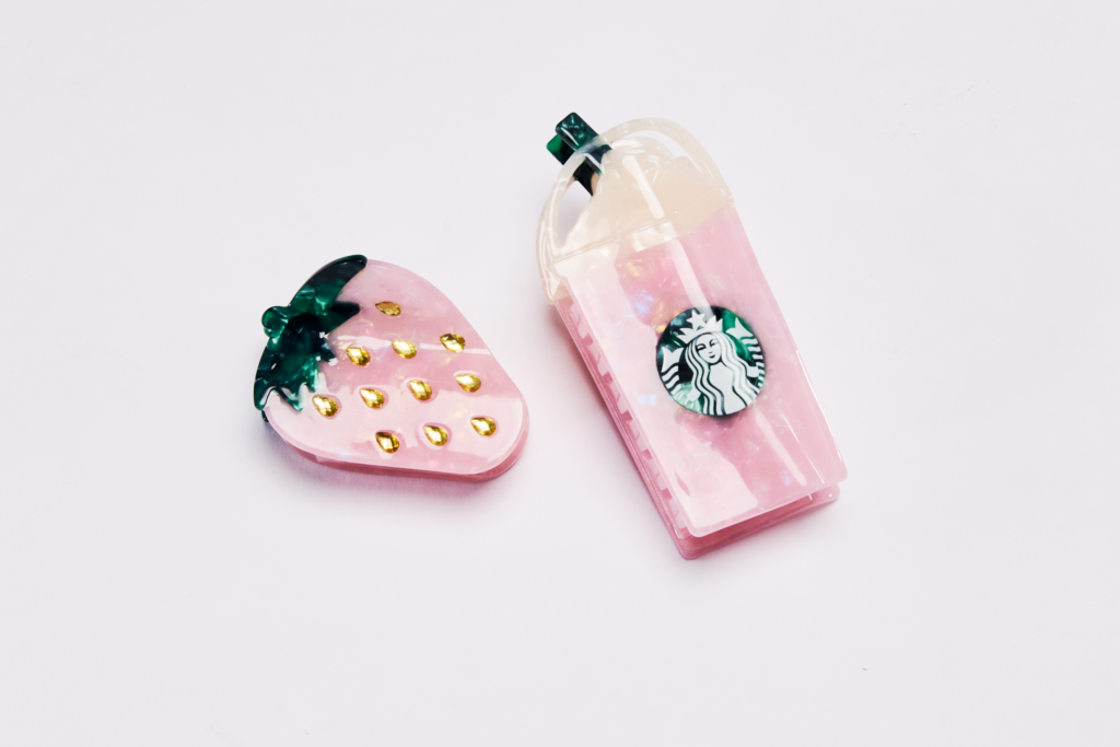 A pink strawberry-shaped accessory with gold accents is next to a pink drink cup resembling a Starbucks frappuccino, featuring a logo.