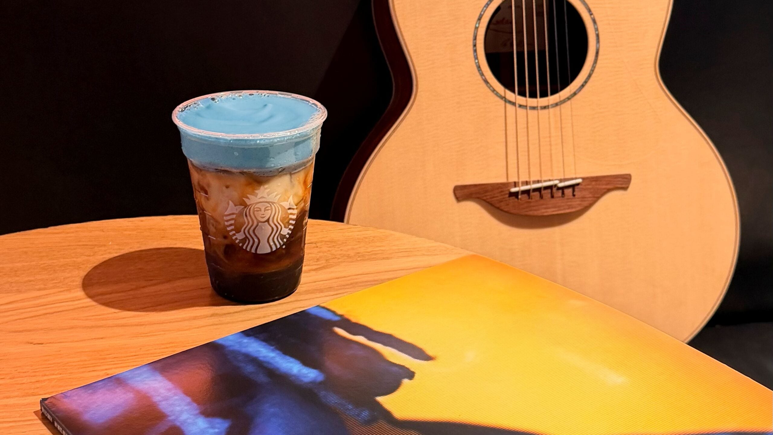 A Starbucks cup with a blue foam topping sits on a wooden table, next to an acoustic guitar and a colorful magazine.