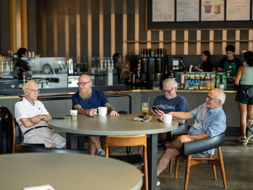 A group of four older men sits around a large, round table in a coffee shop. They are engaged in conversation, with drinks in hand. The background features a coffee counter and menu board.